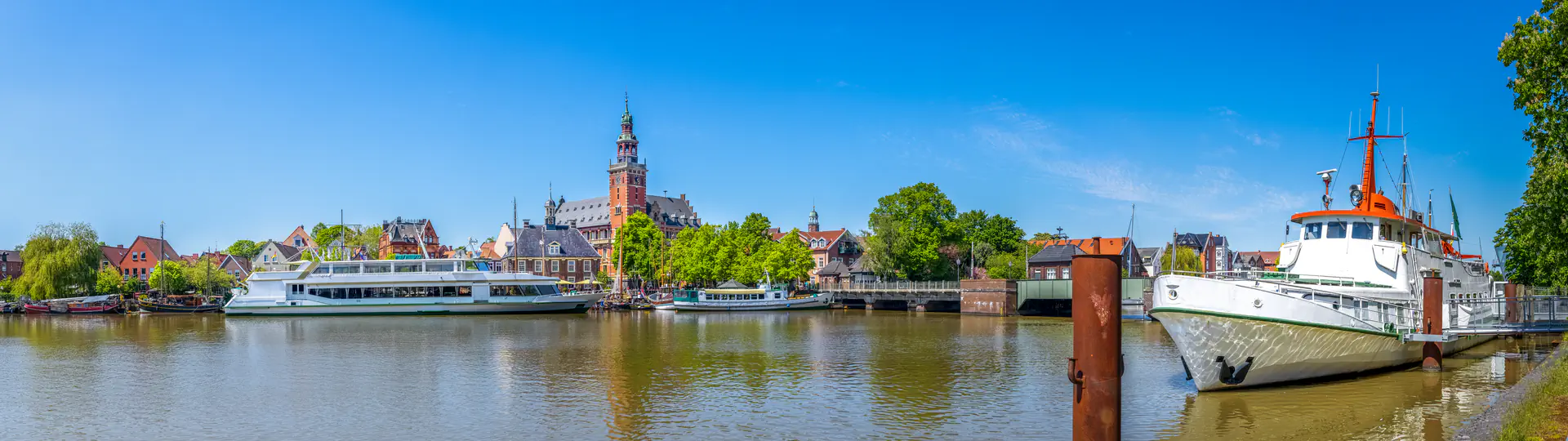 Leer Hafen mit Schiffen, Rathaus und grünen Bäumen unter blauem Himmel.