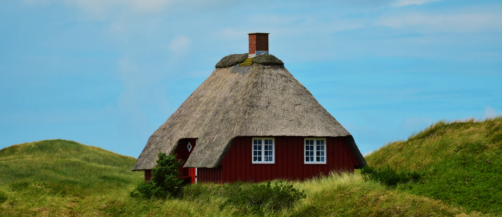 Rotes Reetdachhaus mit Schornstein, umgeben von grünen Dünen und blauem Himmel.