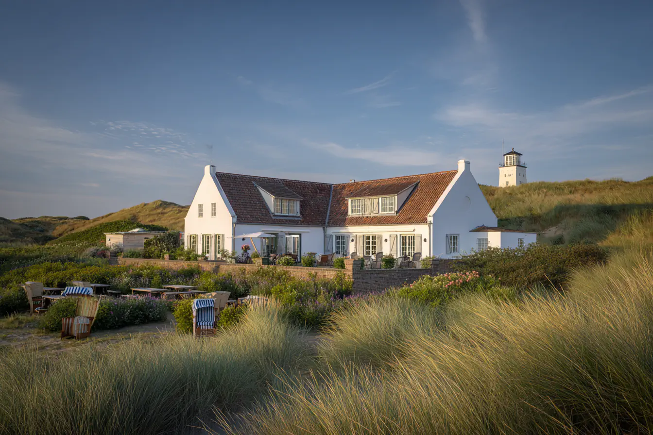 Weißes Haus mit roten Ziegeldach, umgeben von Dünen und einem Leuchtturm im Hintergrund.
